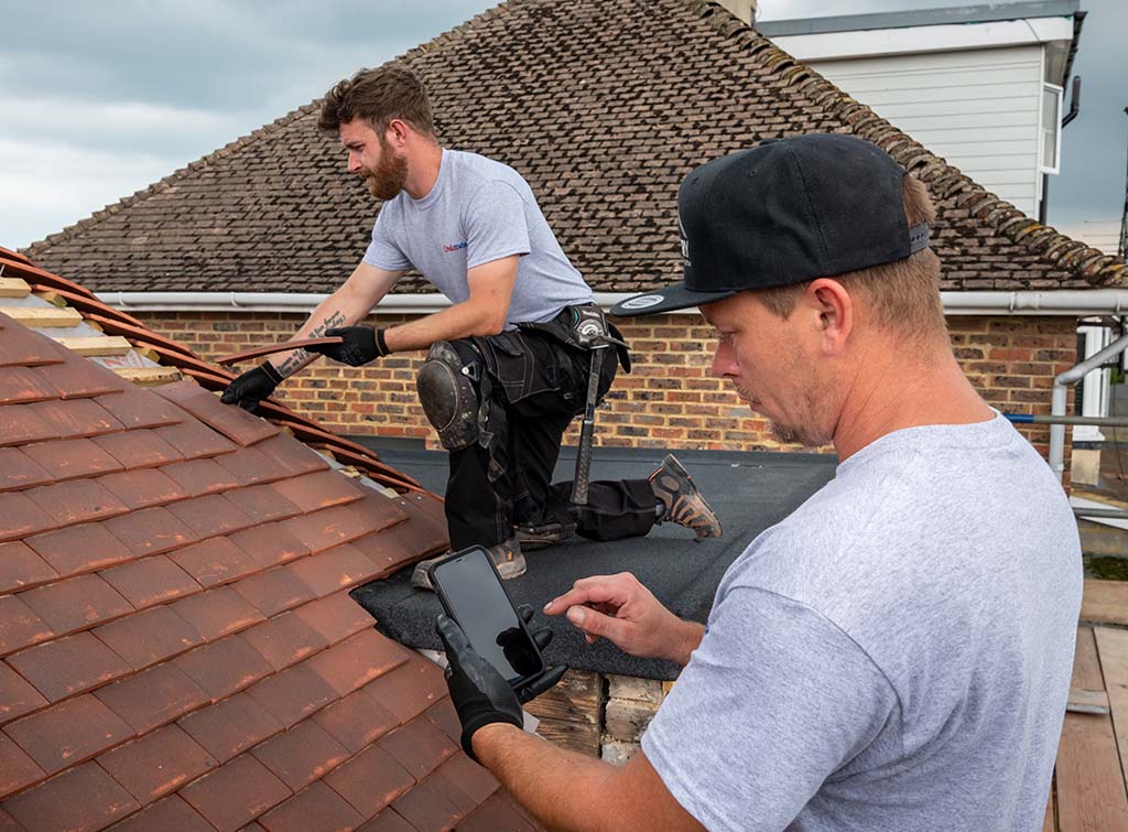 Two Spot Roof Repair roofers on a tile roof, one lifting tiles and one photographing the repair